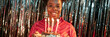 © Seventyfour - Website header shot of cheerful young Black woman smiling at camera and holding birthday cake with lit candles standing against party tinsel backdrop in studio