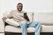 © SHOTPRIME STUDIO - Smiling Black man in a cozy sweater using smartphone on a comfortable couch, showcasing a relaxed atmosphere and modern lifestyle