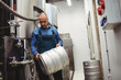 © WavebreakMediaMicro - Male brewer wearing work overalls handling metal beer keg in brewery with fermentation tanks