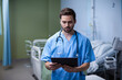 © WavebreakMediaMicro - Male doctor in scrubs standing in hospital with tablet, stethoscope, bed, privacy curtain, IV stand