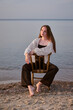 © TRAVELARIUM - Young woman in casual attire sits on chair at serene beach, embodying calmness and introspection. Soft evening light and water create peaceful, reflective mood