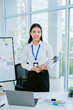 © PSG - Happy Asian businesswoman holding documents in office workspace. Asian female employee using laptop and talking on phone at work.