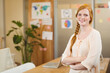 © WavebreakMediaMicro - Red-haired woman in her 20s standing at office table with laptop and corkboard map, copy space