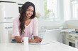 © WavebreakMediaMicro - African American woman typing on silver laptop at white kitchen island near ceramic mug and flowers