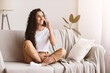 © Prostock-studio - Happy brunette woman with long curly hair having conversation on mobile phone while sitting on couch in living room and looking at copy space. Young lady talking on phone and smiling
