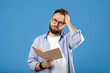 © Prostock-studio - Studying is hard. Young bearded guy having problem understanding textbook on blue studio background. Millennial guy having headache while getting ready for exam or test