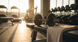 © ON - Studio - Dumbbell rests on bench with a towel, surrounded by gym equipment and weights in a sunlit gym setting. Warm lighting and exercise-focused environment create an atmosphere of fitness motivation