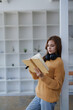 © Jirapong - A young woman in sweater leans against a pillar reading a book indoors.