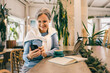 © kostikovanata - Senior woman with gray hair using smartphone in cafe, with laptop, coffee and notebook on the table