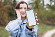 © sav_an_dreas - Smiling young woman showing blank smartphone screen to camera outdoors.