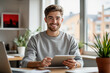 © f_bossa - A young man with light brown hair and a beard smiles warmly while holding a tablet in a modern, sunlit office setting.