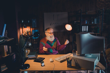 elderly male programmer working on computer in home office illuminated by warm light