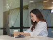 © Oil And Den - A young woman wearing glasses and a white shirt sits working on a laptop outside a glass building.