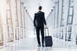 © Prostock-studio - Rear view of tall businessman in stylish suit walking by airport terminal, carrying luggage, copy space. Unrecognizable entrepreneur arrived at airport, having business conference abroad
