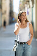 © JackF - Young female tourist around 20 years old with wavy long brown hair walking through European city in summer, casually wearing blue jeans and white top, looking natural and unposed with slight smile