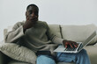 © SHOTPRIME STUDIO - Thoughtful young African American man working on a laptop in a cozy living room, wearing a knitted sweater, expressing concern or contemplation