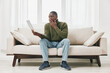 © SHOTPRIME STUDIO - Concerned African man sitting on a sofa with a laptop, showing surprise and anxiety in a minimalist home setting with bright natural light