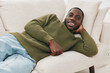 © SHOTPRIME STUDIO - Young Black man in green sweater relaxing on a sofa, smiling joyfully, showcasing comfort and happiness in a cozy indoor setting