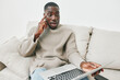 © SHOTPRIME STUDIO - Young African man in cozy sweater looking surprised while sitting on a sofa with a laptop, expressing emotions of shock while working from home