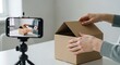 © Richafuji - A person is opening a cardboard box on a white table with a smartphone recording the process. The scene takes place indoors with natural light.