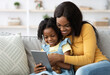 © Prostock-studio - Technologies For Leisure. Happy Black Mother And Little Daughter Using Digital Tablet Together While Sitting On Couch At Home, African American Mom And Female Child Shopping Online With Modern Gadget