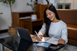 © Apichat - A woman is sitting at a table with a laptop and a notebook