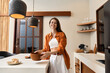 © SHOTPRIME STUDIO - Young woman in an orange shirt and white pants smiles while holding a bowl in a modern kitchen, conveying happiness and home cooking