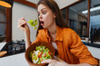 © SHOTPRIME STUDIO - Surprised young woman with long hair in casual orange shirt, holding a bowl of fresh salad, showcasing healthy eating in a modern kitchen setting