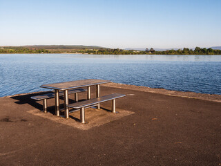 Wall Mural - Picnic area with metal table and sit on a pier with stunning view on surrounding nature scene with lake and blue cloudy sky. Barbeque spot in a public place. Nobody.