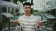 © Krakenimages.com - Young hispanic man on terrace holding phone looks contemplative outside a city cafe wearing striped shirt surrounded by tables and chairs in an urban outdoor setting