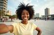 © miss irine - Happy black woman takes selfie smiling in city park. Smiling female with afro hairstyle poses for photo outdoors, enjoying sunny day. Content creator takes profile picture for social media.