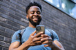 © Dejan - A young man happily interacting with his smartphone, showcasing joy and engagement in an urban setting with a brick wall backdrop.