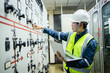 © Sumala - Electrical engineer is performing inspection in substation control room with control panel while holding laptop and wearing safety gear for precise monitoring and control