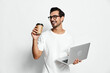 © SHOTPRIME STUDIO - Cheerful smiling young man wearing glasses holding coffee cup and laptop while standing against plain backdrop. Modern lifestyle and work concept.