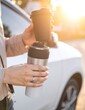 © Natty - A woman in casual attire stands beside her parked car, joyfully raising her coffee cup, sunlight glinting off the vehicle's surface, creating a warm atmosphere.