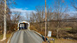 © Greg Kelton - A historic covered bridge spans over a small rural road. The structure is framed by bare trees, with a clear blue sky overhead. A weight limit sign indicates three-ton restrictions for vehicles.