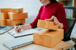 © PSG - Businesswoman sitting to check customer orders and pack boxes to ship online at home. Small business entrepreneur, SME, freelancer, online business, work from home.