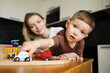 © Seluna - Little boy playing with a toy police car on a wooden train track, looking curiously at the camera while his smiling mother watches from behind.