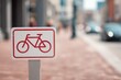 © RICH - Red-white bicycle lane sign with bicycle icon, brick-red sidewalk and blurred urban street background conveying clear directional guidance.