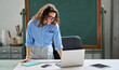 © insta_photos - Young busy happy woman school professional teacher standing at desk in front of chalkboard in classroom working on laptop computer. Online tutor teaching remote education virtual classes.