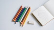 © sattaya - An overhead view of a group of colored pencils, an open sketchbook with a pencil drawing, and an eraser, all arranged on a white surface.