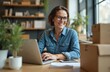 © Pete - Smiling business woman in office using laptop. 40 years old entrepreneur working online. Female owner wearing denim shirt, glasses, sitting at desk. Boxes, tablet, plants, shelves background. Modern