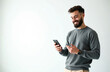 © Pete - Young bearded man in grey sweater uses smartphone. Smiling guy holds device, watches, chats online, browses social media, uses apps. Modern lifestyle, tech, communication.