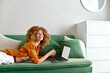 © SHOTPRIME STUDIO - Young woman with curly hair working on a laptop at home, wearing an orange shirt and white shorts, exuding a joyful emotion in a cozy, green themed living room