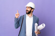 © luismolinero - Young architect man with helmet and holding blueprints over isolated purple background thinking an idea pointing the finger up