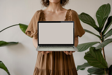 Wall Mural - Woman in Earthy-Toned Dress Presenting Laptop with Blank Screen, Cozy and Natural Setting