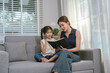 © Witoon - An Asian mother and daughter sit together indoors, smiling and bonding over a book, enjoying digital learning and joyful family time.