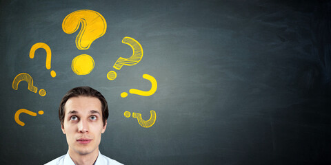 Fototapete - Thoughtful man looking up at yellow question marks on blackboard background with copy space for education or problem-solving concept.