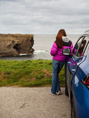 Wall Mural - A woman in a purple jacket stands next to a blue car in a parking lot and looking at a stunning nature scene with cliff, rough stone coastline and cloudy sky. Travel and tourism. West of Ireland.