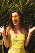 © zvkate - Excited young woman in yellow floral dress and yellow sunglasses smiling outdoors on a sunny day, standing in front of palm leaves with joyful expression. Here am i!